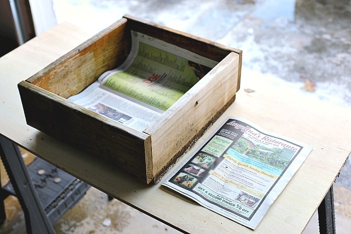 wooden box with newspaper on the bottom. The box and newspaper on resting on top of a wooden table. There is also a stack of newspaper next to the box
