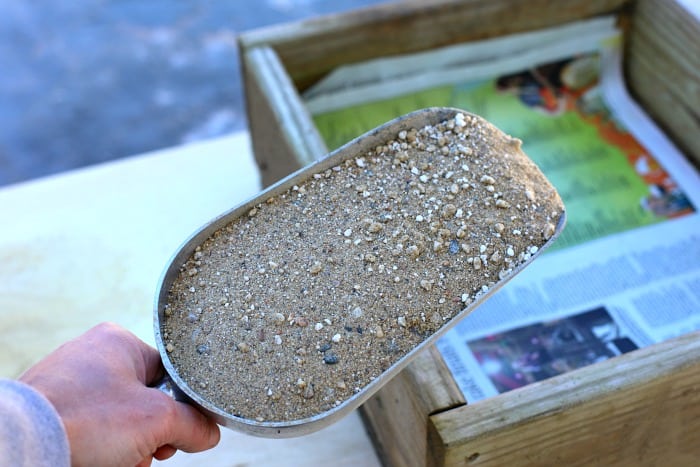 a hand holding a silver scooper filled with sand and perlite getting ready dump into wooden box lined with newpaper
