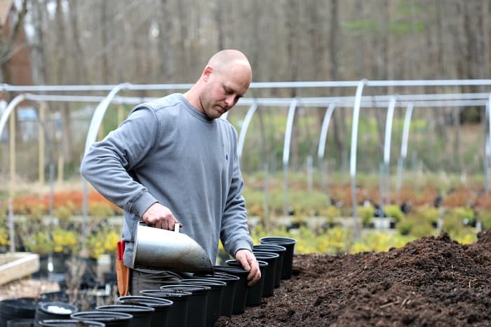 man holding silver scooper and filling black pots with potting mix